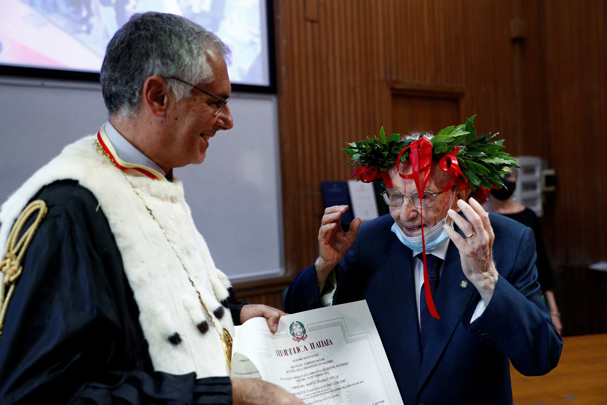 An old man with a suit wearing a crown of laurels smiles while another man, slightly younger and dressed as a college dean hands him a graduation certificate