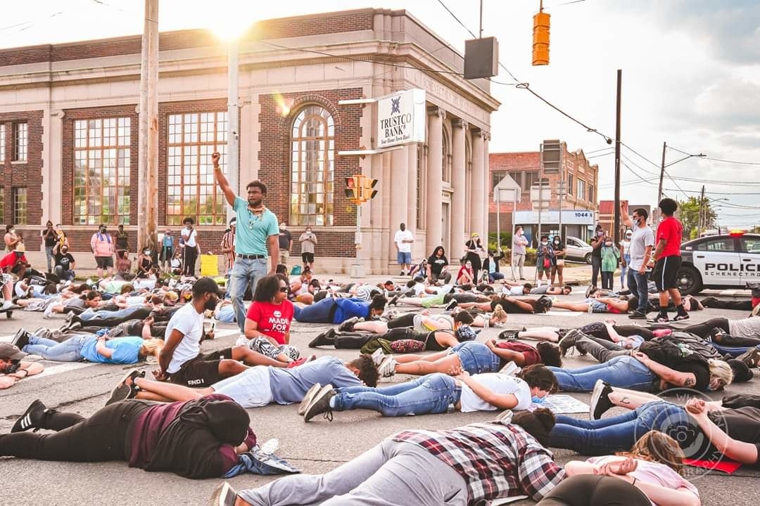 One man stands in the middle of the street with his arm raised while people lay face-down with their arms behind their backs on the ground all around him