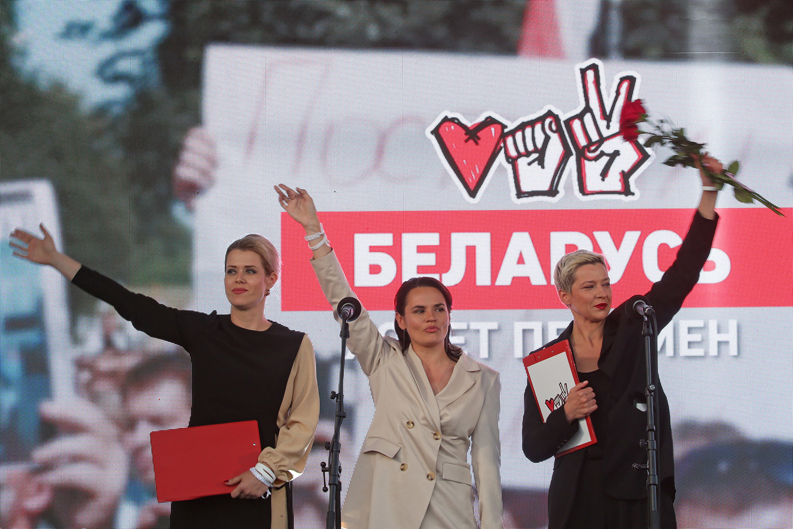 Three women stand on a stage with the arms raised, waving to the crowd. Behind them are their campaign symbols: a heart, a fist, and a hand making a &quot;V&quot; for victory