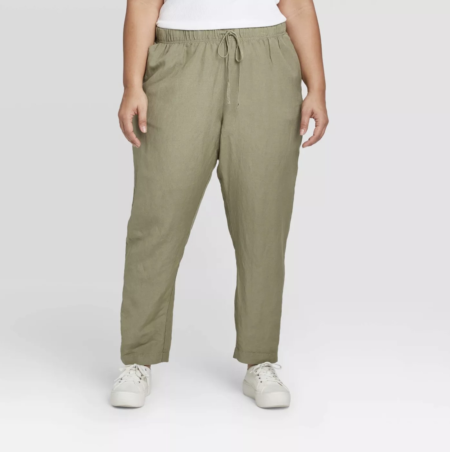 Model is wearing olive green linen skinny ankle pants with a drawstring waist and white tennis shoes
