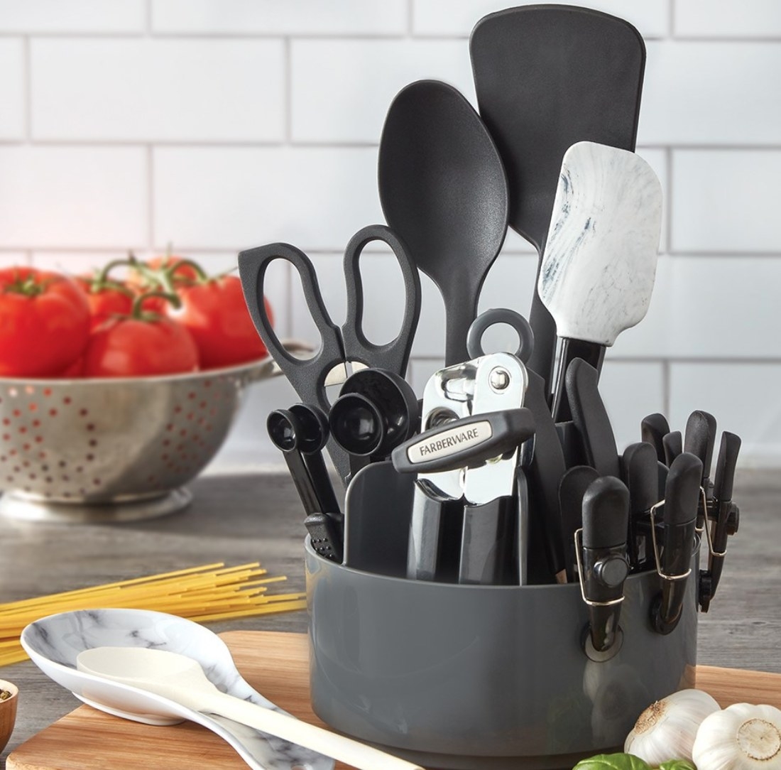 A rotating caddy filled with 18 cooking utensils in front of a white tile wall and bowl of tomatoes