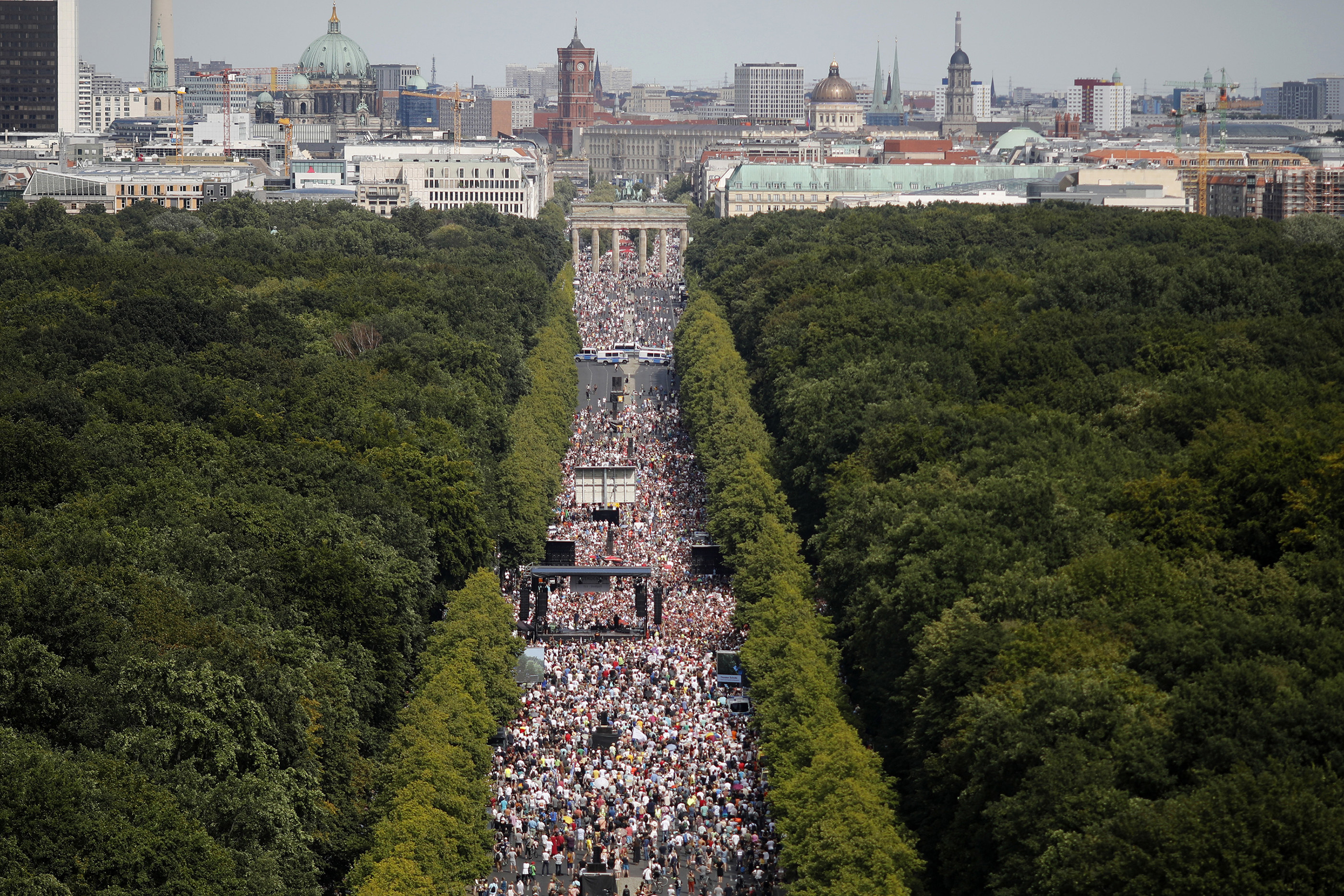 An aerial shot of thousands of people walking down a boulevard surrounded by trees and toward the skyline of Berlin 