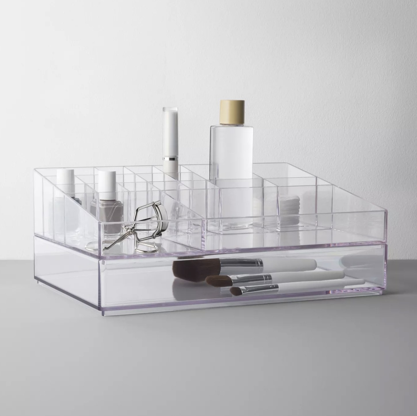 A clear cosmetics organizer holding nail polish, makeup brushes, a bottle of mascara, a few cotton pads, a lash curler, and a bottle filled with a clear solution