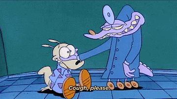 Rocko sits on the ground while a purple animal dressed as a doctor holds Rocko&#x27;s eyes. The doctor says &quot;Cough, please&quot;