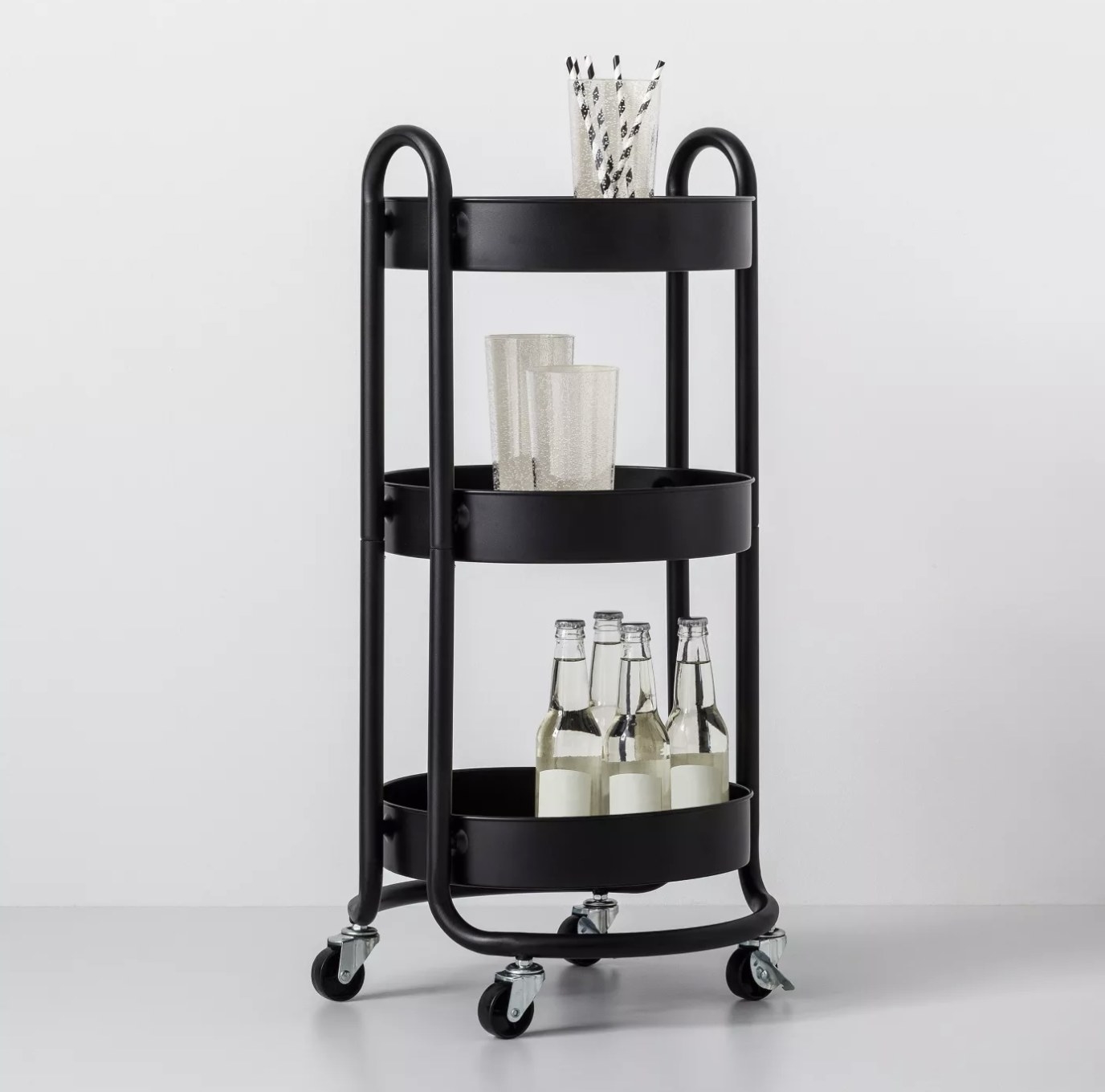 A black cart with casters and three shelves holding glass bottles, cups, and a cup of straws