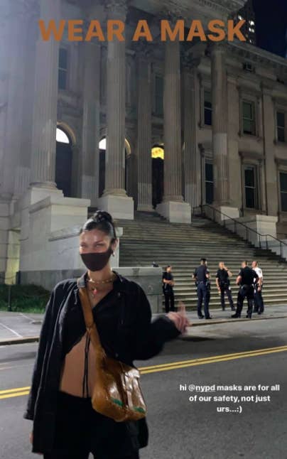 Bella Hadid wearing a mask posing for a photo while flipping off the camera with unmasked officers behind her