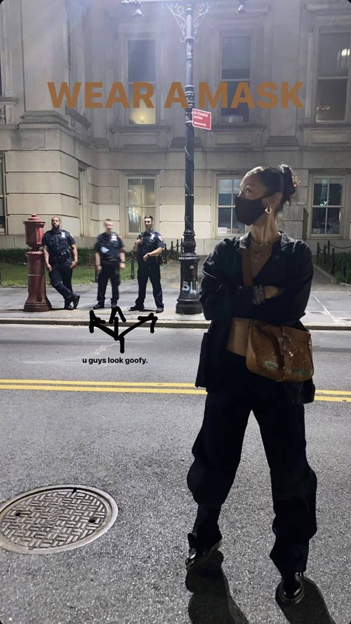 Bella Hadid standing on a street at night with her arms crossed wearing a mask, with three unmasked officers behind her in the distance