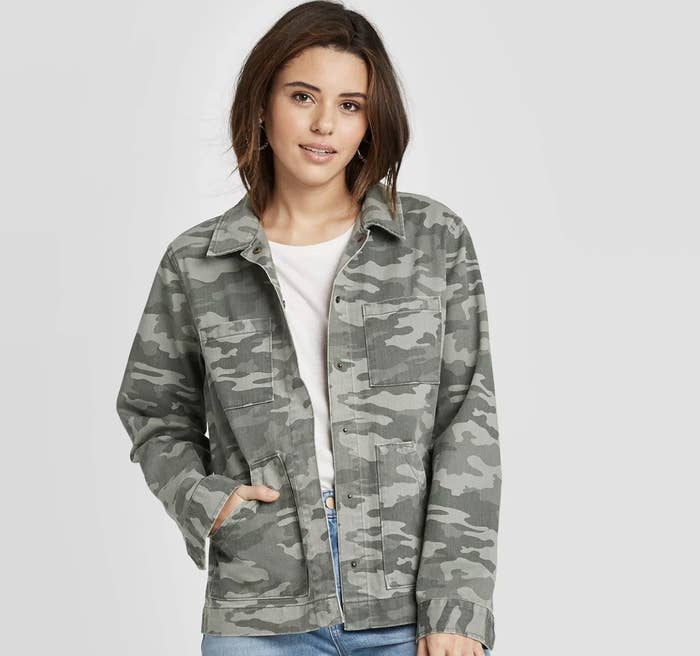 Model wears camo print jacket with white tee and jeans. 