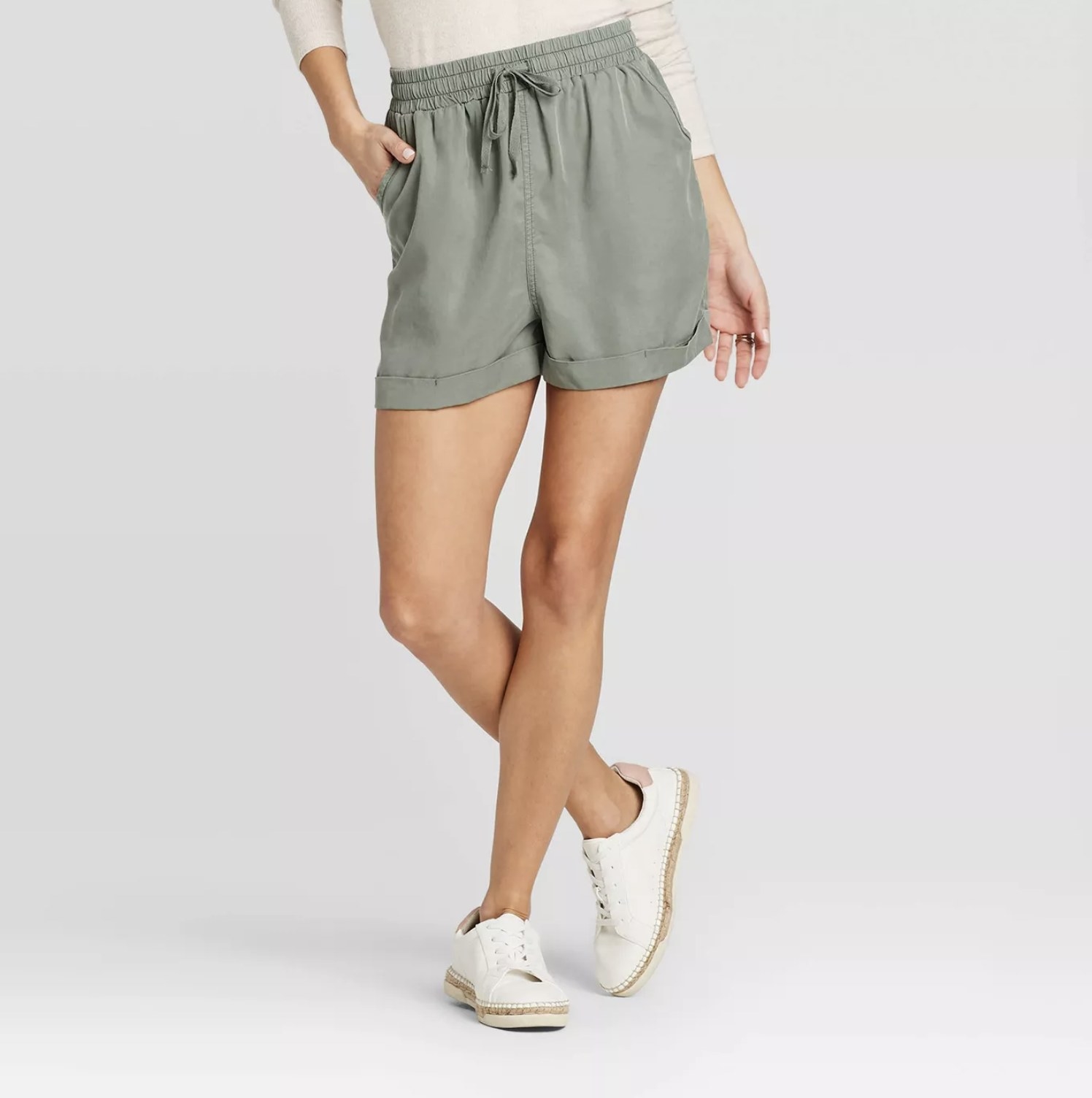 Model wears olive green shorts with white long sleeve tee and white sneakers. 