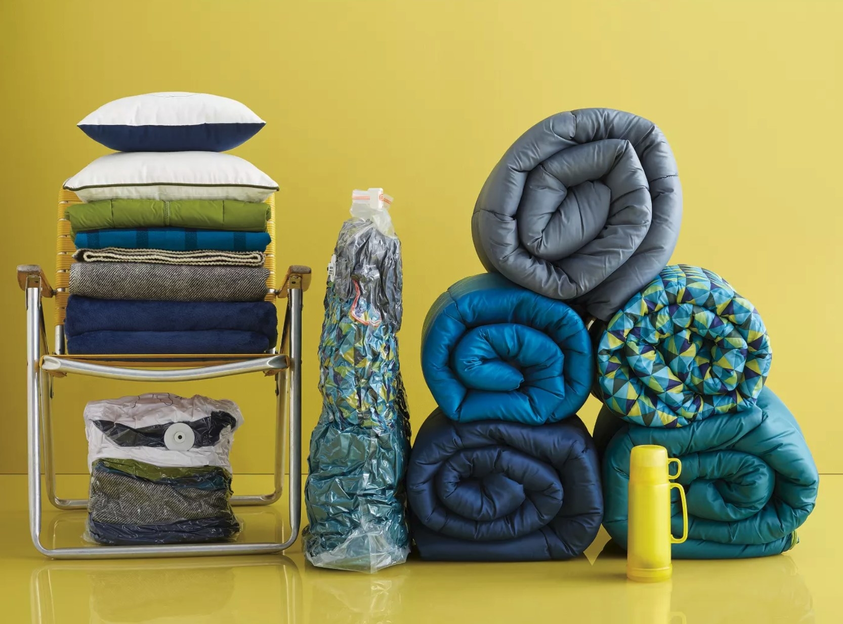 A stack of rolled-up sleeping bags next to a compressed bag of three sleeping bags and a stack of blankets and pillows in a chair above a compressed bag of a matching stack of blankets and pillows.