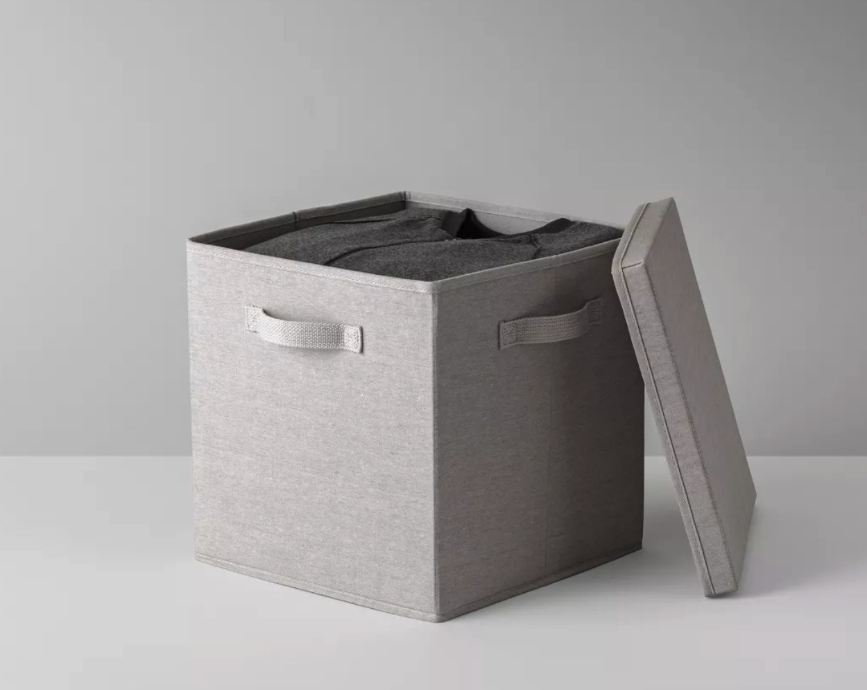 A gray, fabric storage cube with handles with the lid off to reveal a dark gray zip-up.