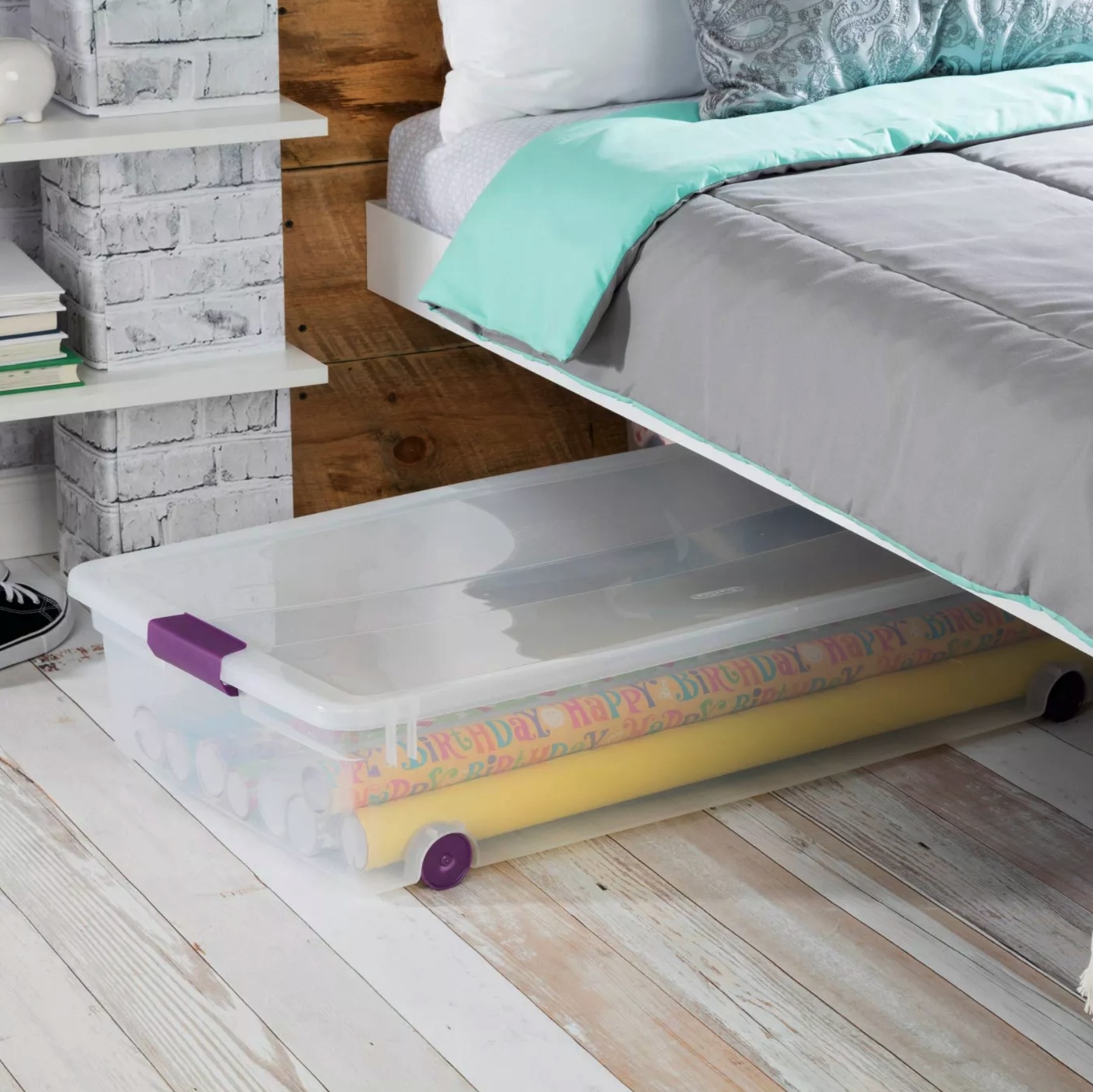 A long, clear plastic bin with purple wheels and latches halfway under a bed. It is holding several rolls of wrapping paper.