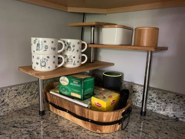 Reviewer pic of the three-tier shelves with wood tops and silver legs in the corner of a counter with mugs and assorted other products on it and below it