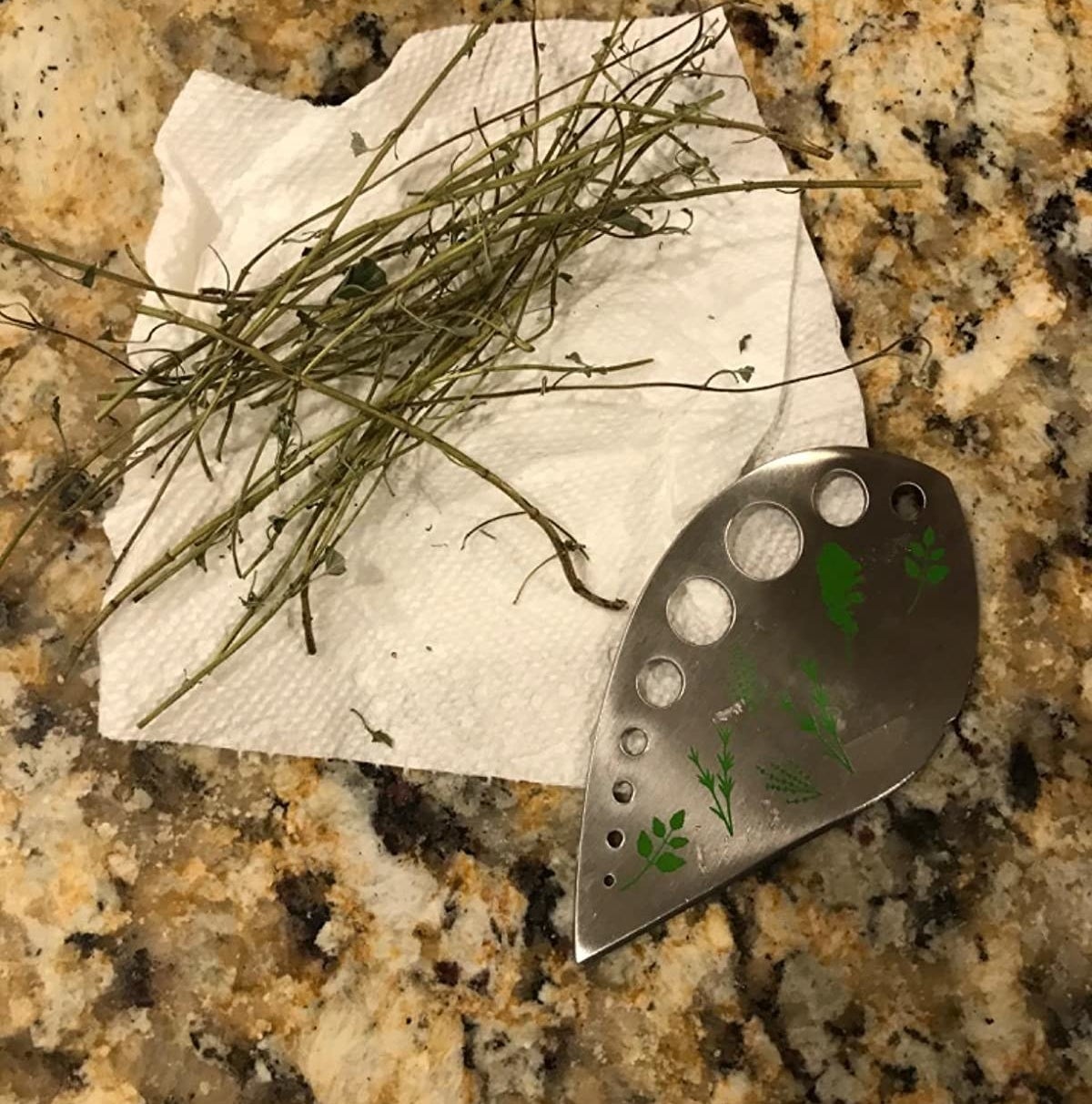 reviewer's teardrop-shaped herb stripper with holes featuring illustrations of what herbs to strip where on one side and a sharp edge on the other side nest to stripped rosemary stalks with few remaining leaves