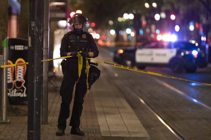 A police officer wearing a helmet ties tape reading "POLICE TAPE DO NOT CROSS" around the crime scene while a squad car flashes its sirens in distant background