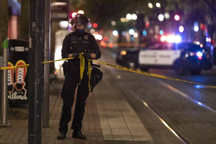 A police officer wearing a helmet ties tape reading &quot;POLICE TAPE DO NOT CROSS&quot; around the crime scene while a squad car flashes its sirens in distant background