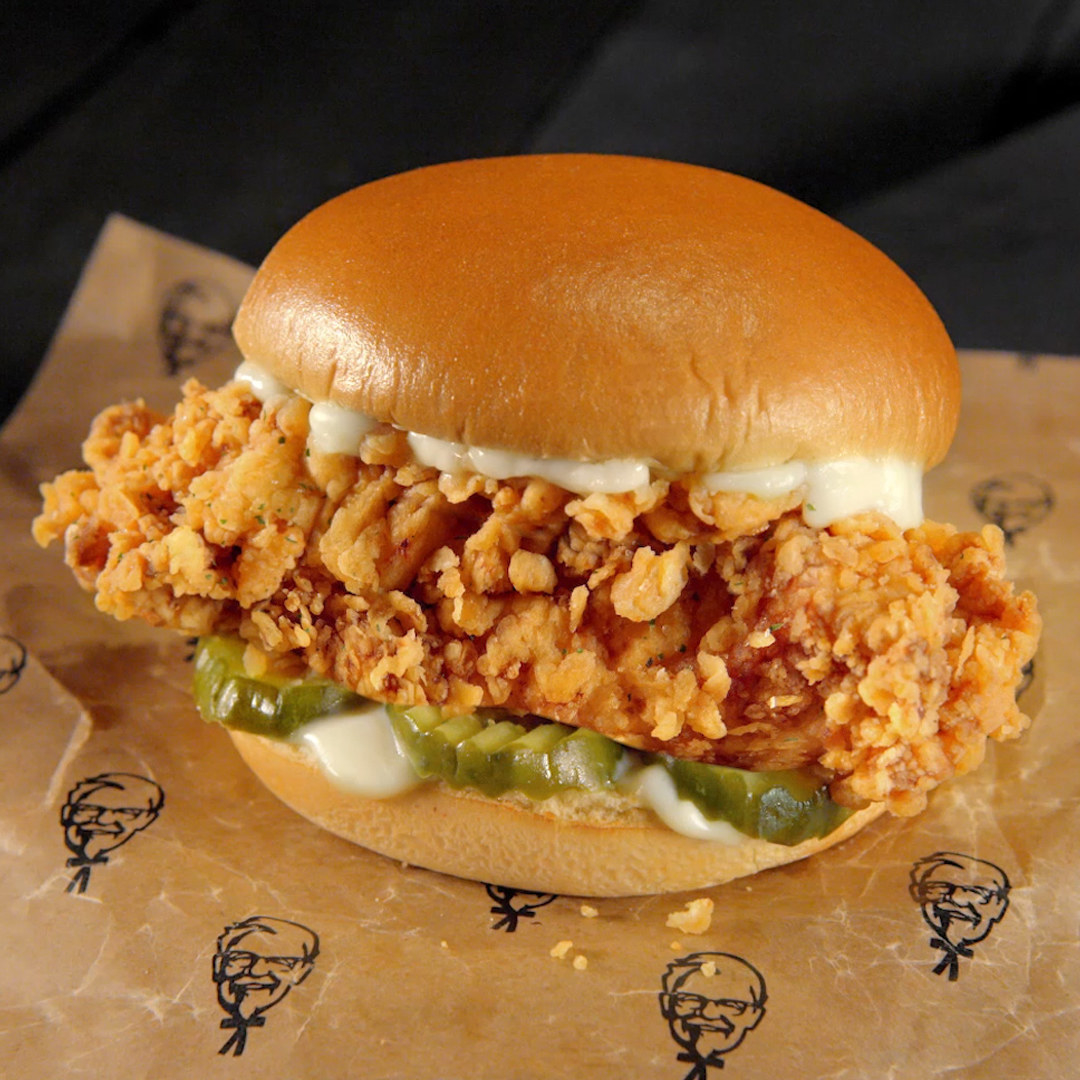 The Famous Chicken Chicken Sandwich is on top of parchment paper that has Colonel Sanders&#x27;s illustrated head on it. The sandwich is against a black background.