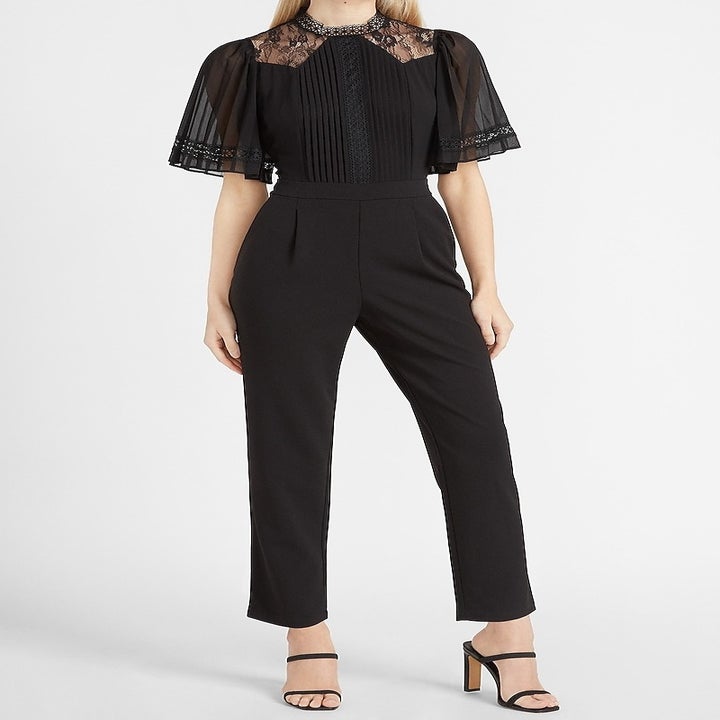 A model in the black jumpsuit with short sheer sleeves, a lace panel at the neck, and a pleated front