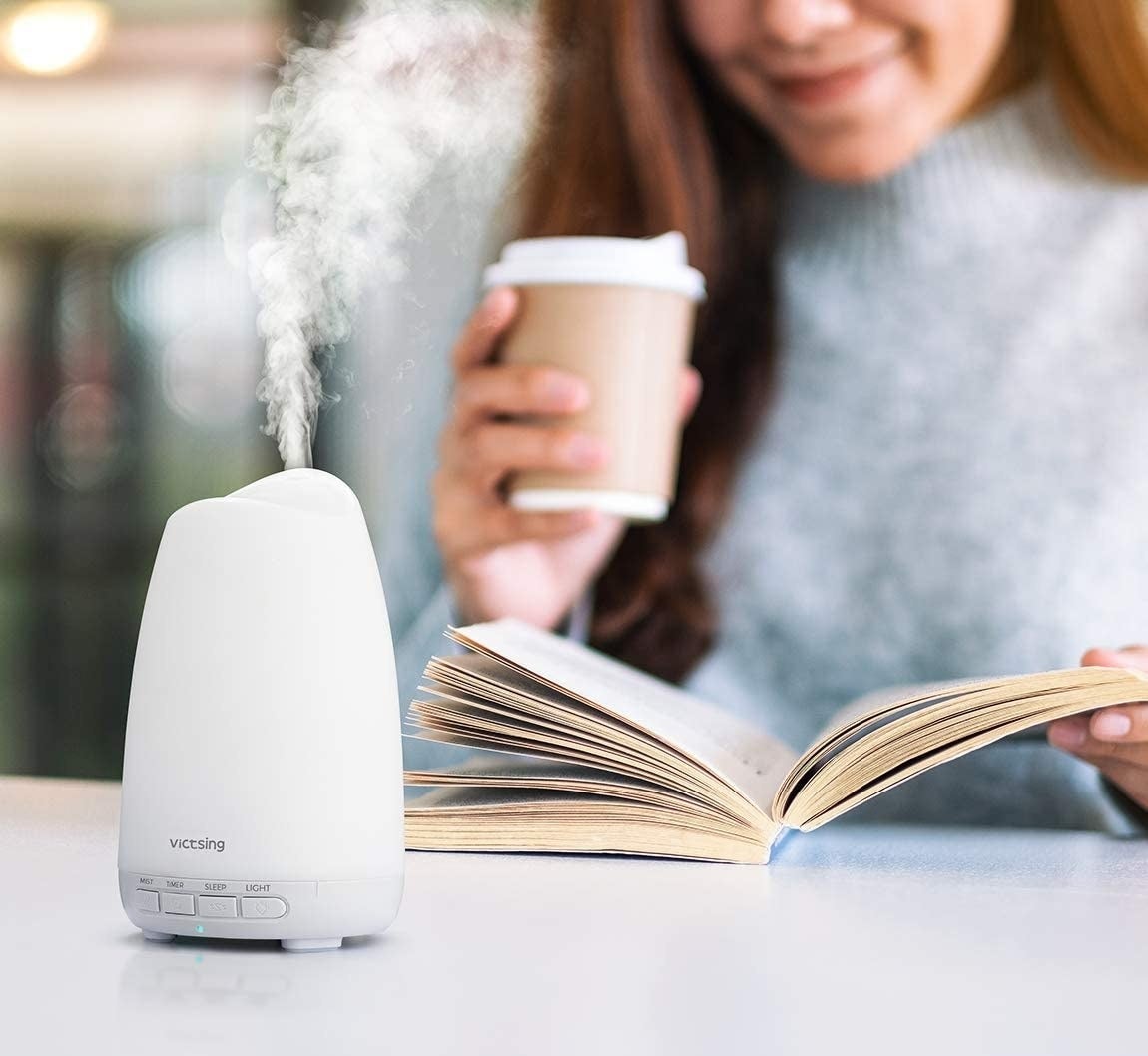 A person drinks a coffee while reading a book with the diffuser running in the foreground