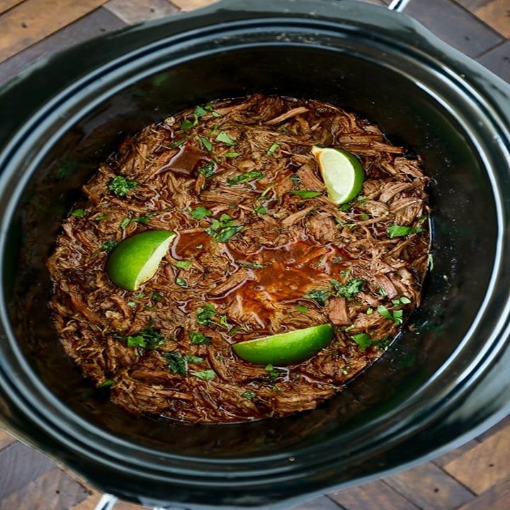 Shredded barbacoa beef in a slow cooker with fresh herbs and lemon wedges.