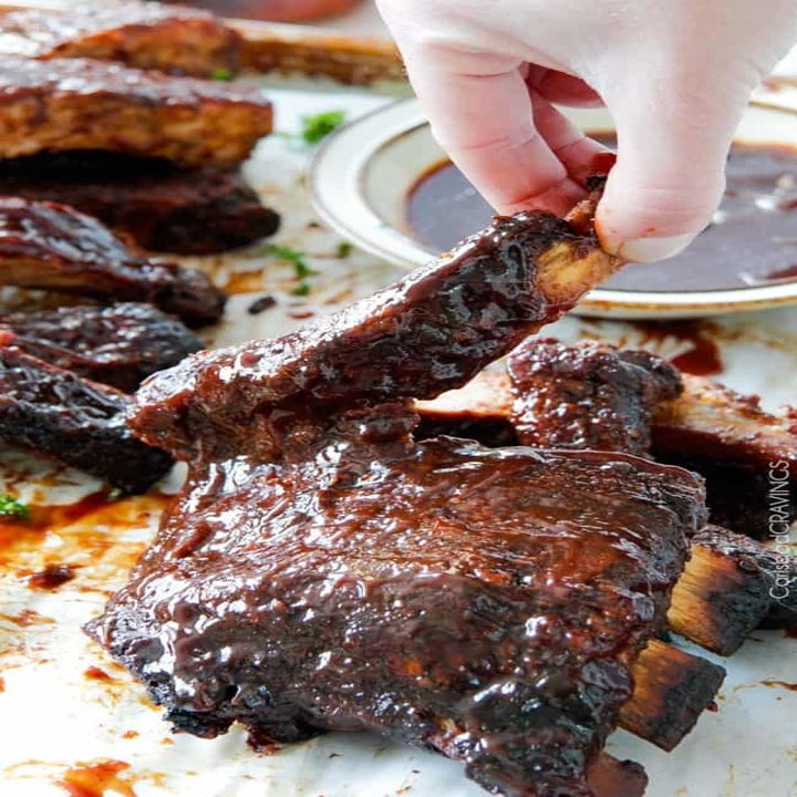 A hand holding a sticky, glazed rib above a whole rack of ribs.