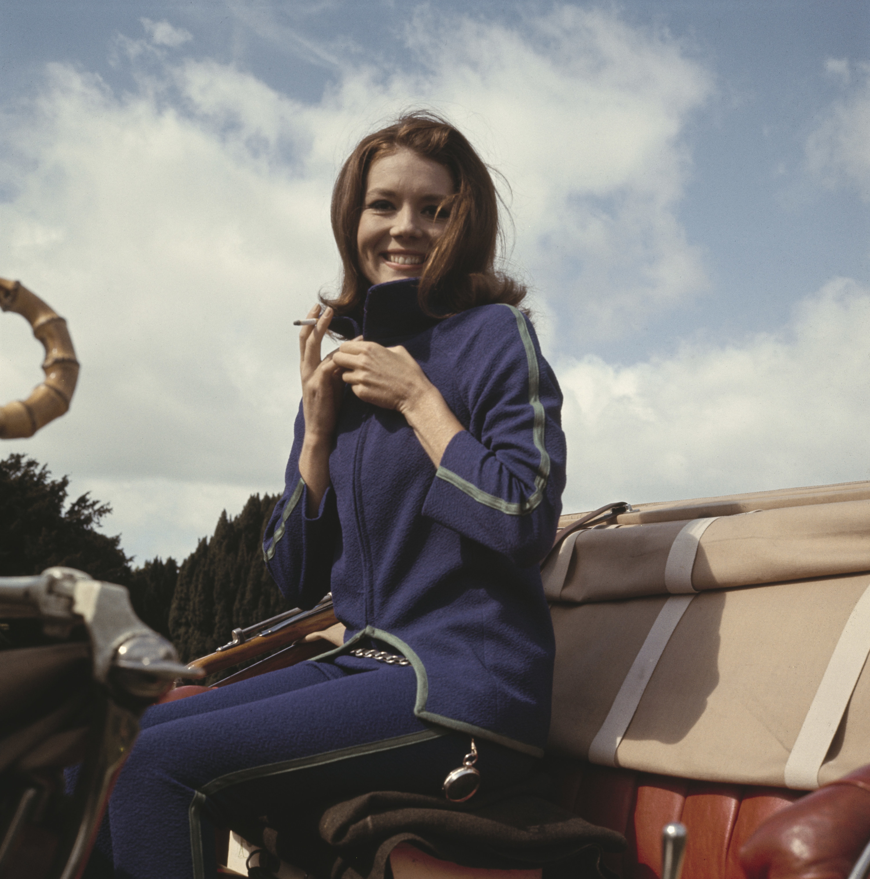 Rigg, wearing a blue tracksuit with green trim, holding a cigarette while seating in the driver&#x27;s seat of a car during filming of British television series The Avengers