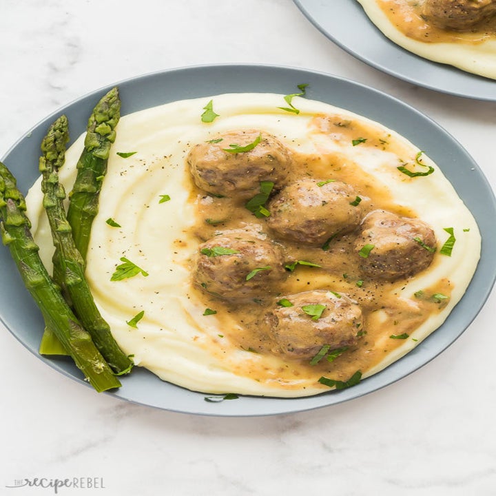 A bowl of Swedish meatballs over mashed potatoes with asparagus on the side.