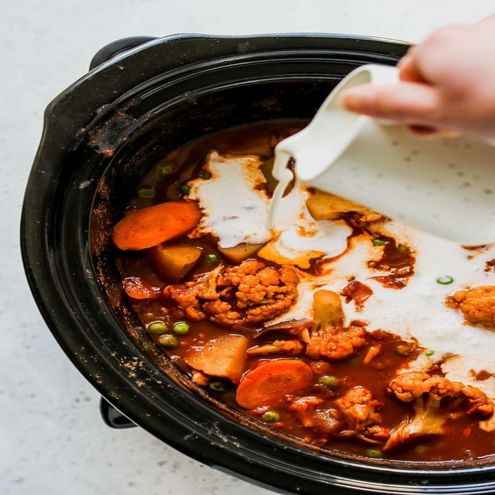 Pouring coconut milk into a slow cooker filled with cauliflower, carrots, peas, and spices.