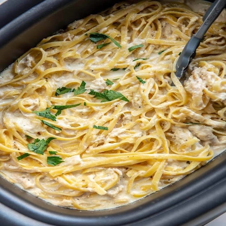 A slow cooker filled with long spaghetti noodles in a cream sauce with shredded chicken.