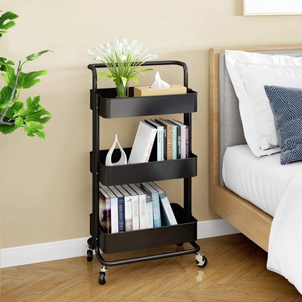 A metal rolling cart with three tiers Two shelves have books and the top shelf has a box of tissue and a vase with flowers