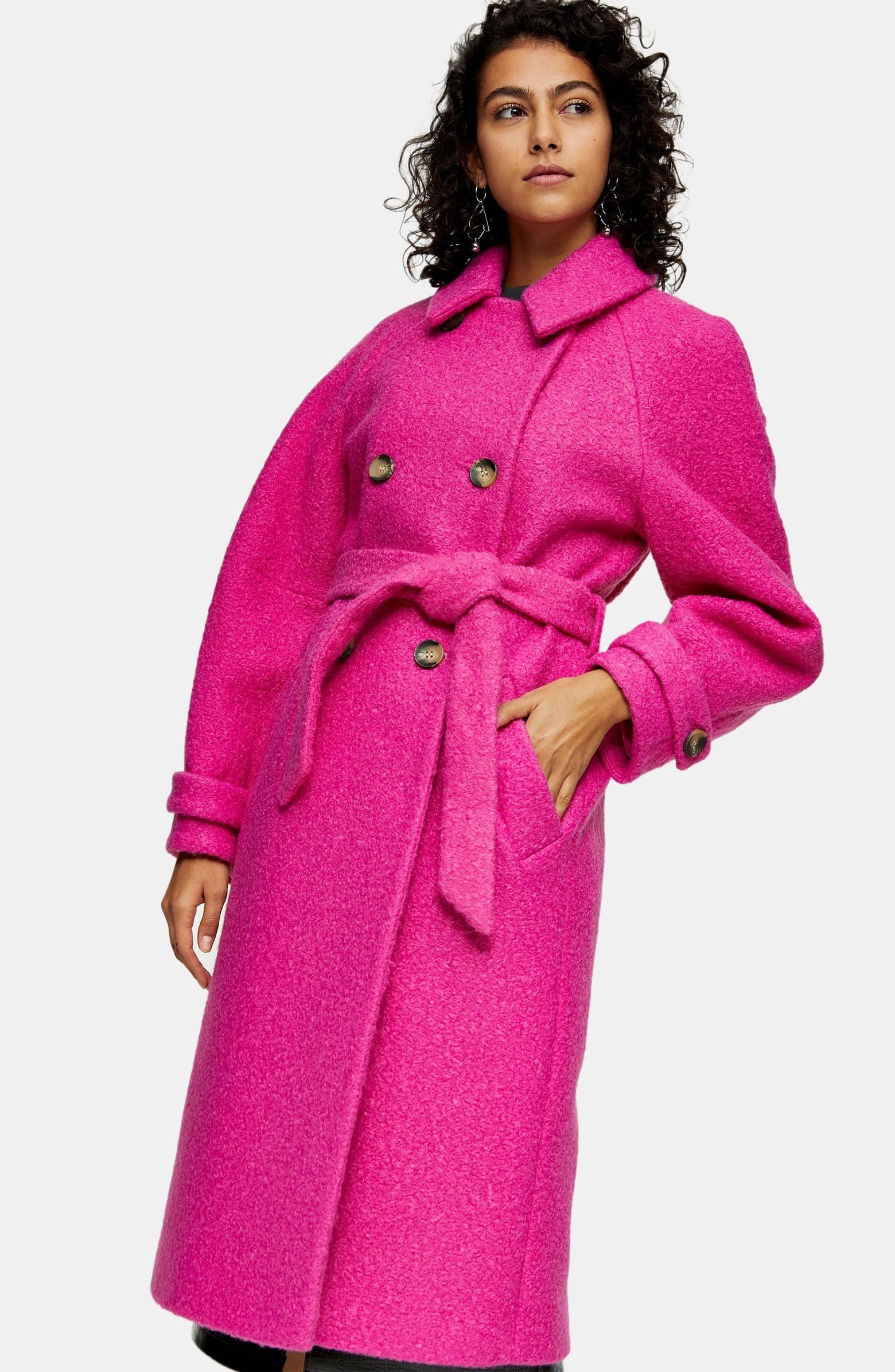 model wearing a long wool-like coat that's a magenta pink with a wrap-around belt, pockets, and double-breasted buttoning