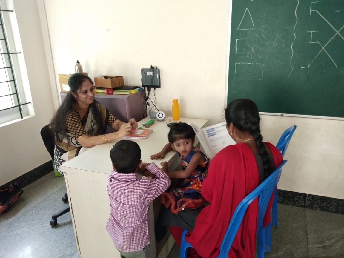 a mother, with a baby on her lap and the teacher smiling at her, looks at her kid&#x27;s report card during a parent teacher meeting