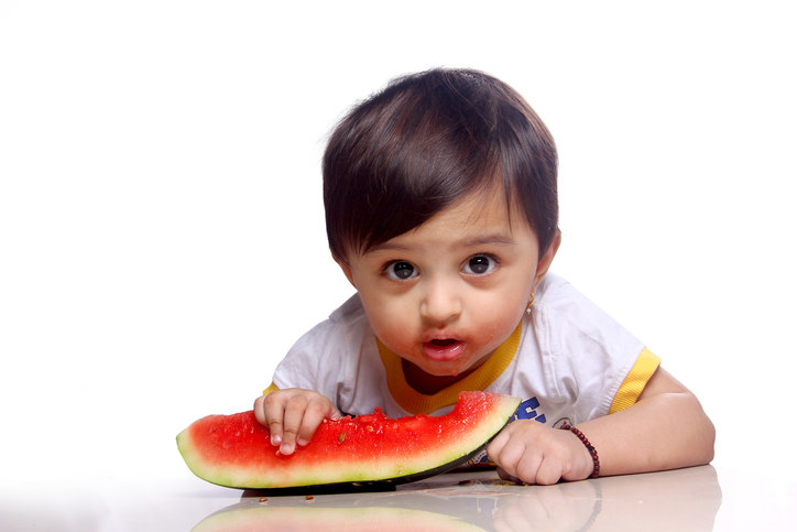 A small baby stares into the camera while eating a watermelon slice