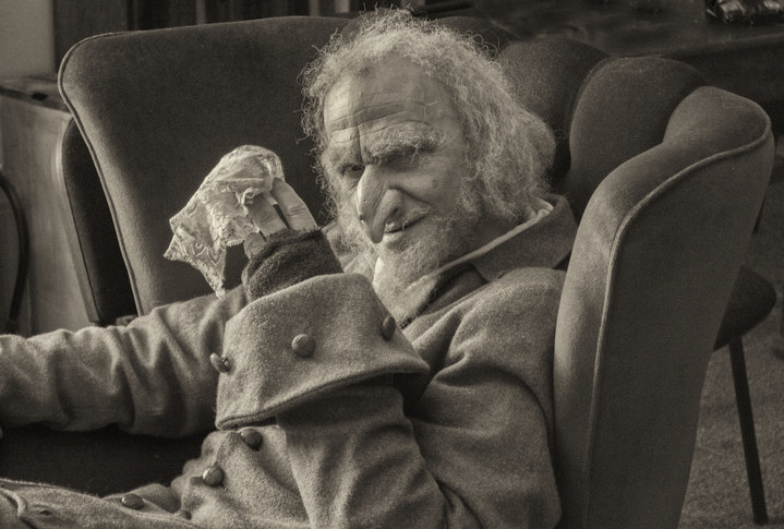 Black and white image of a mature man with long white hair, wearing scruffy clothes sitting in an old chair holding a white handkerchief