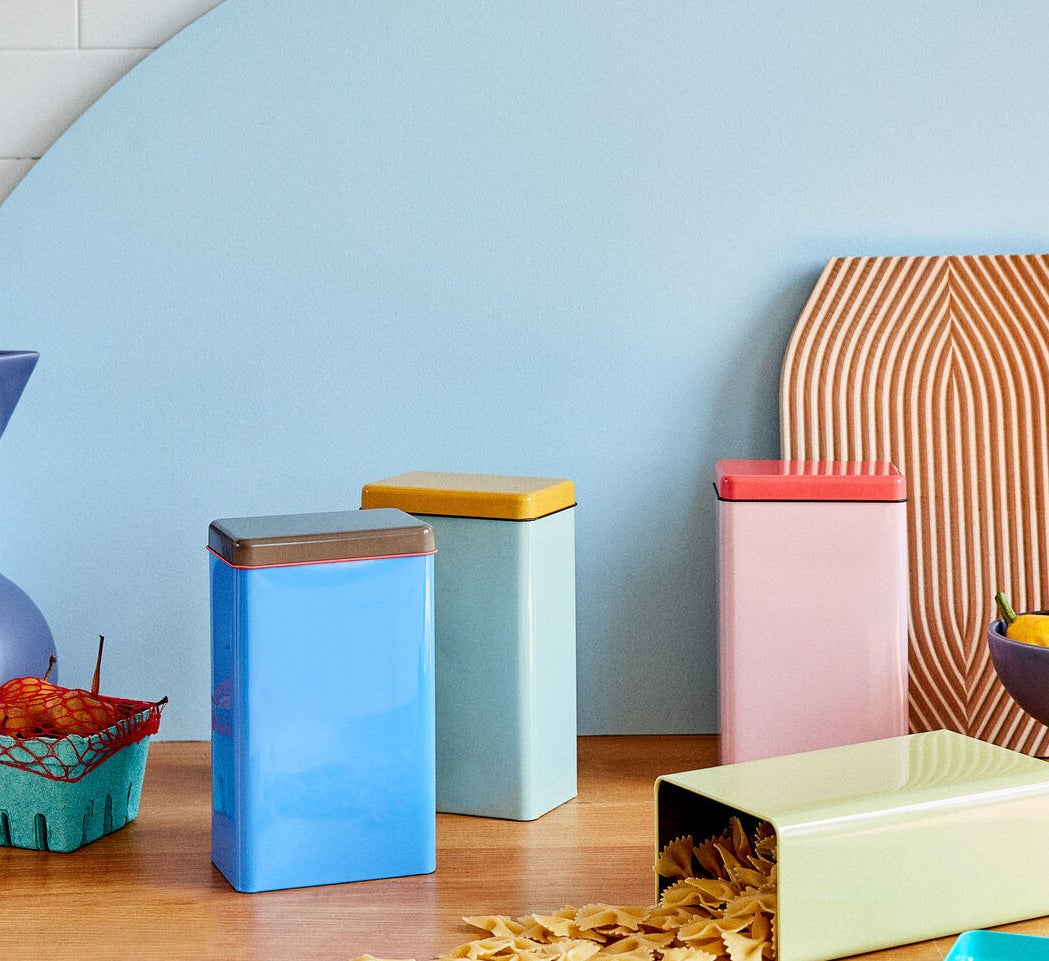 yellow, pink, blue, and green rectangular tins with lids in a darker color with dry pasta in one