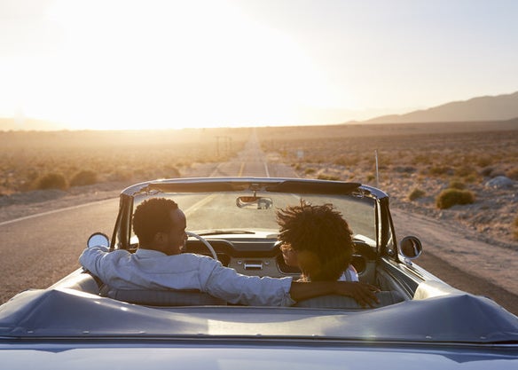 A couple smiles at each other in a convertible as it drives down an open road 