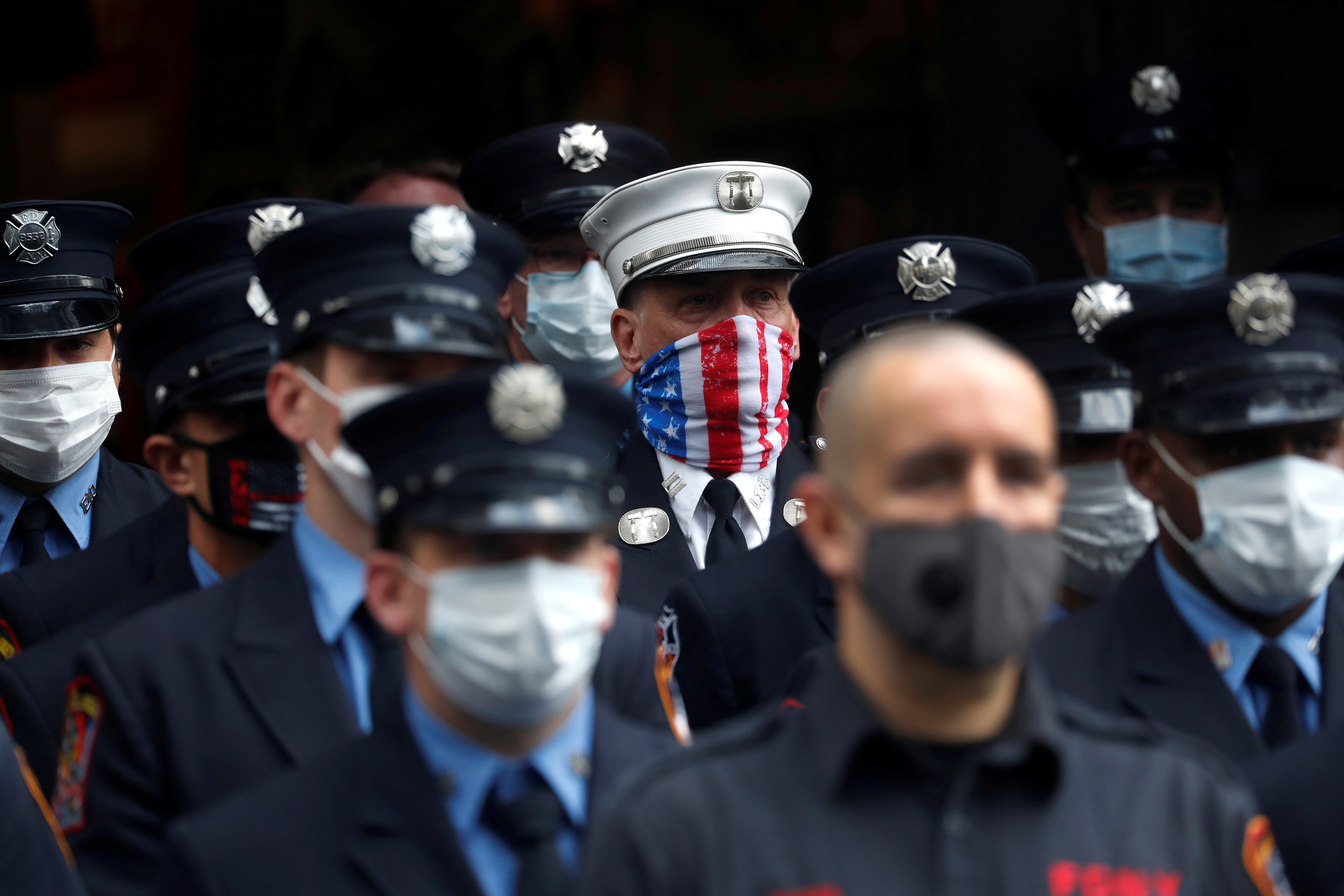 Several uniformed firefighters stand in a row wearing white or gray face masks; in the background one man wears a face mask with a US flag print