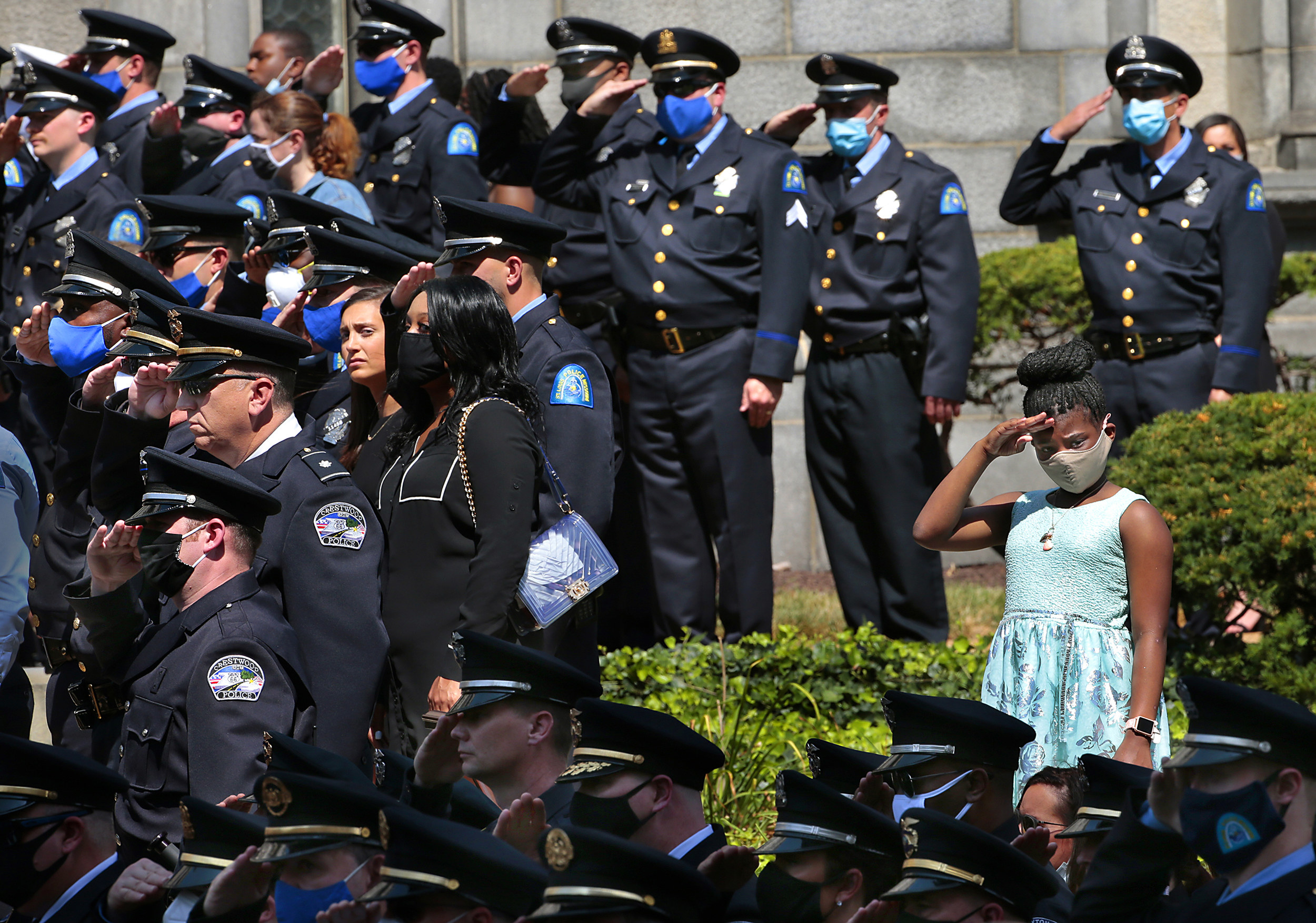 A young girl wearing a face mask and light blue dress salutes with several dozen uniformed police officers around her