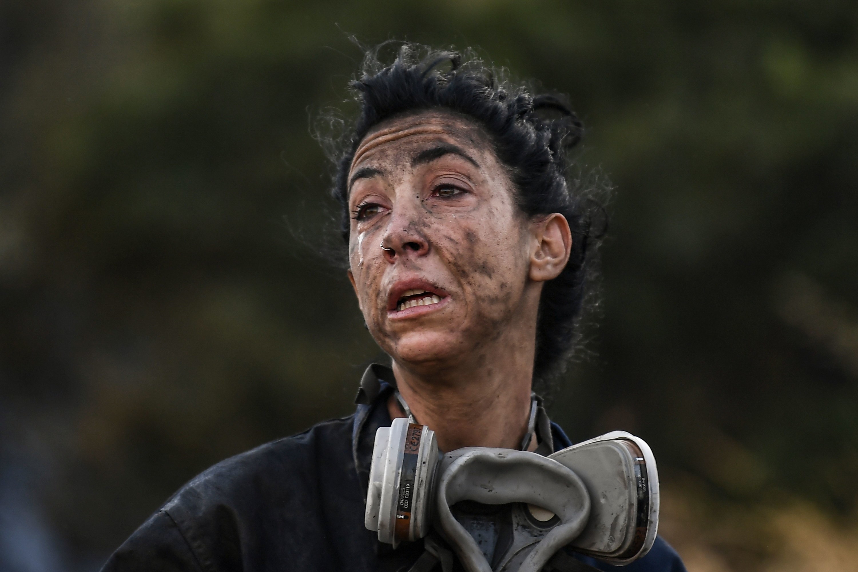 A firefighter with an ash-covered face and a breathing apparatus around her neck grimaces