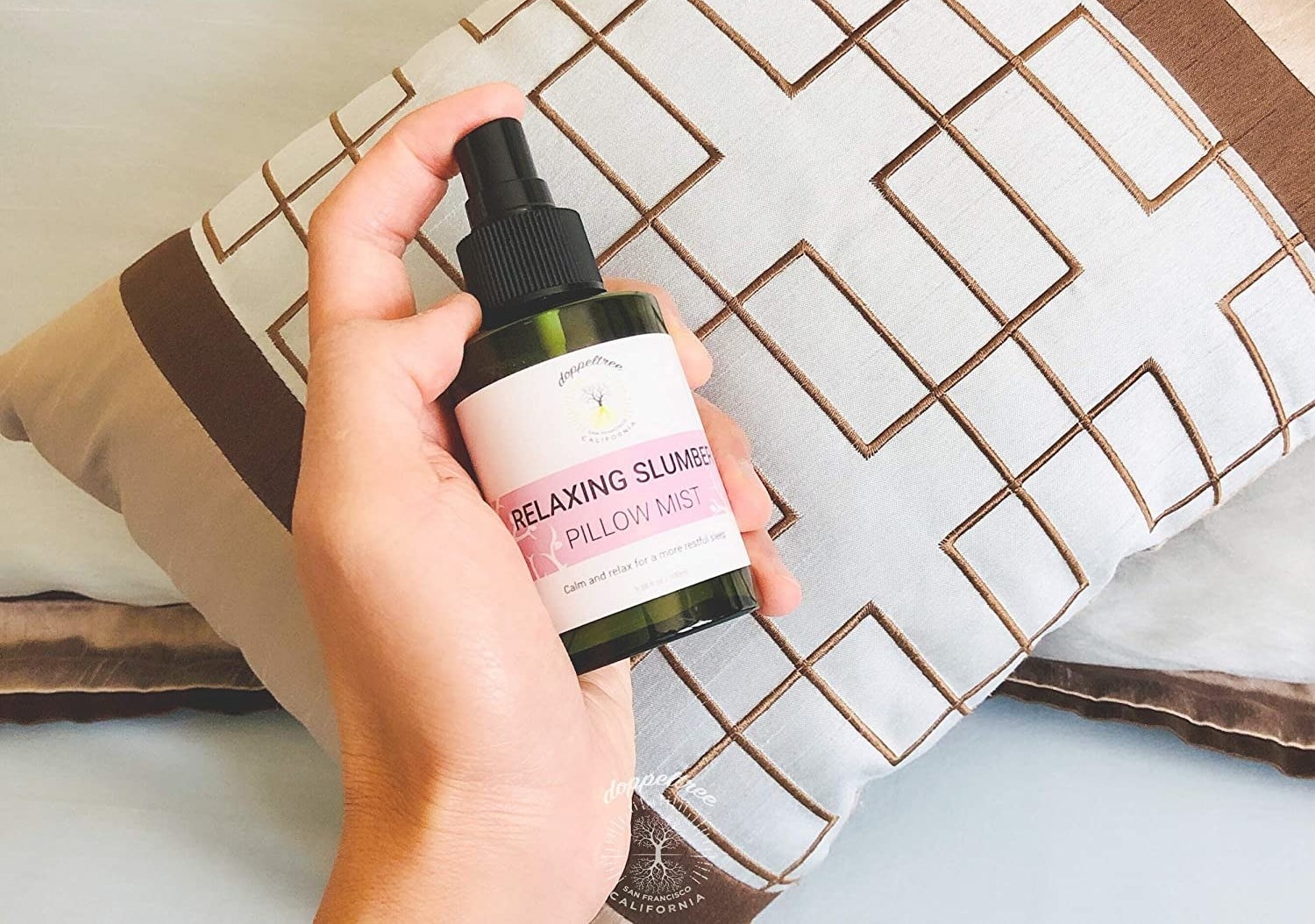 A hand holds a bottle of the product on a pillow