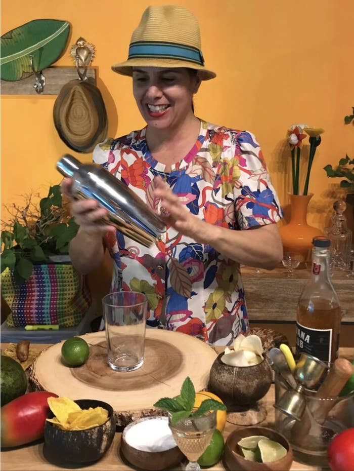 A woman in colorful clothes smiles and shakes a cocktail shaker as she&#x27;s surrounded by  cocktail ingredients