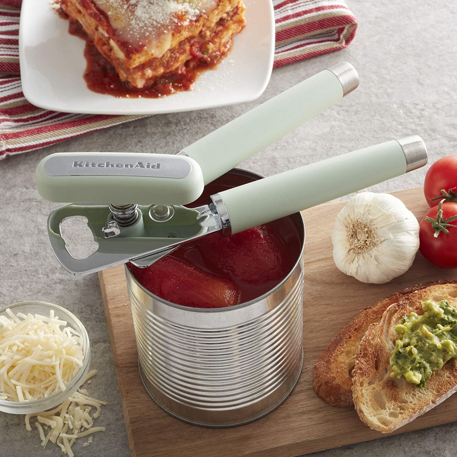 An open can of tomatoes next to some cheese, garlic, and toast with the can opener perched on top