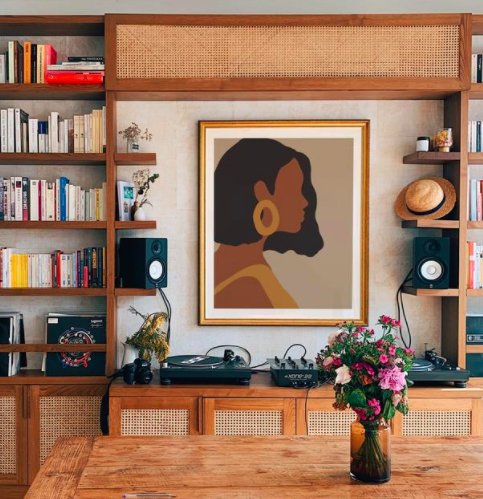 Framed art print of African American woman wearing gold hoops and a yellow dress on a wall about a wooden bookcase and DJ equipment