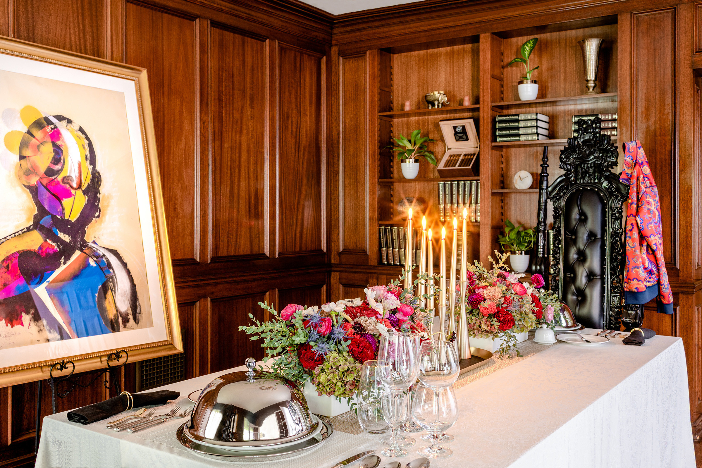 A tableclothed table topped with flowers, crystal glass, candles, and covered silver platters with an ornate, tall black chair at the head