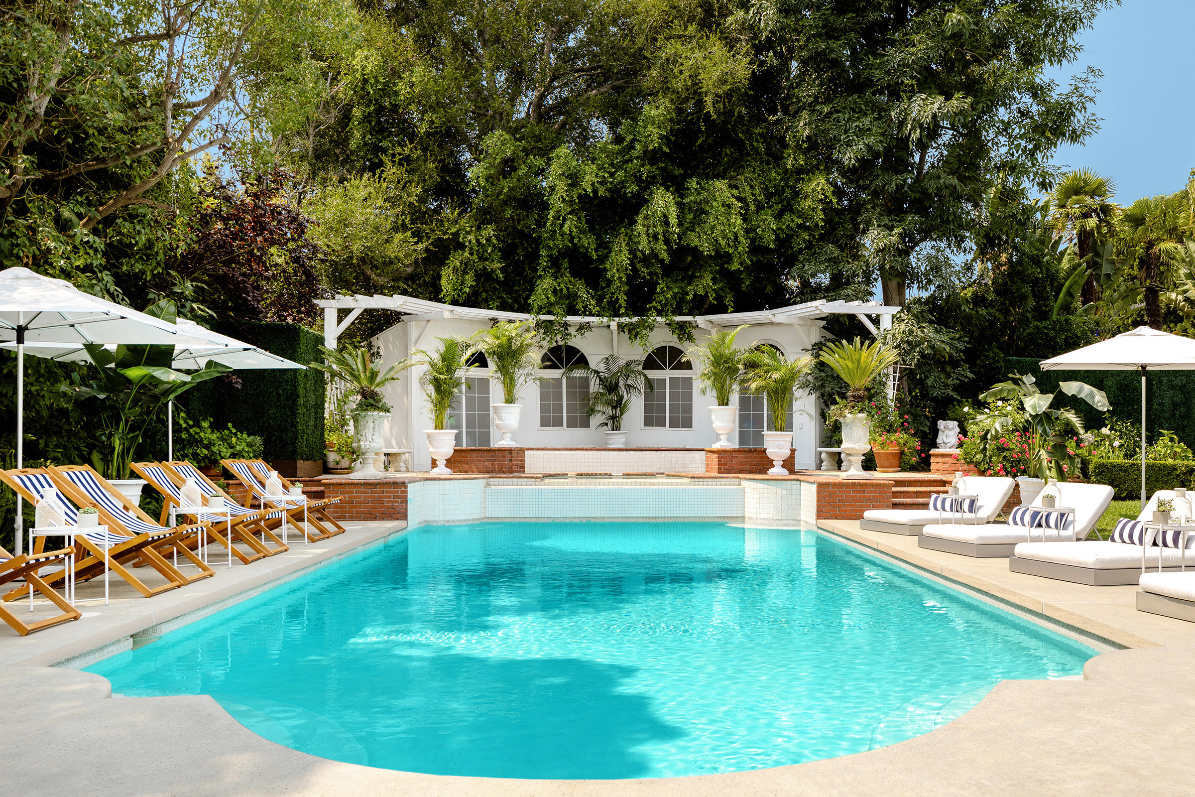 A wide shot of a turquoise pool surrounded by lounge chairs, umbrellas, and a backdrop of lush trees