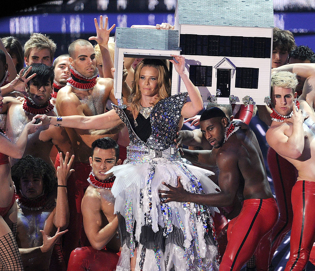 Chelsea Handler on stage wearing a doll house on her head and surrounded by shirtless back-up dancers