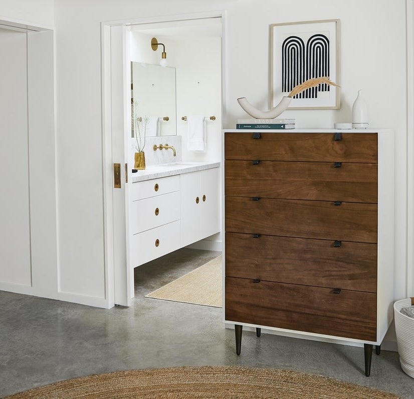 The wood five-drawer dresser with leather pull tabs and white frame with dark brown legs in a room. 