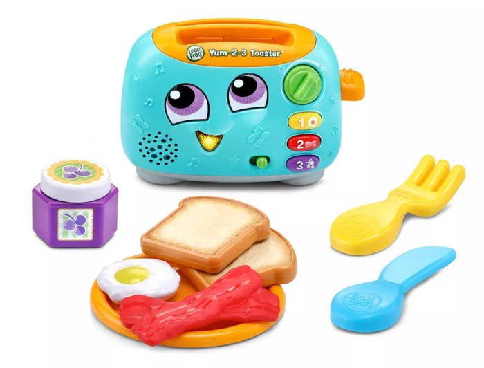 Teal and orange plastic toaster toy with a face along with a plate of toy food and utensils