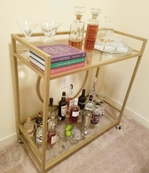 Reviewer pic of the bar cart with two glass-top shelves with a geometric pattern to them and assorted alcohol bottles and glasses on each one.