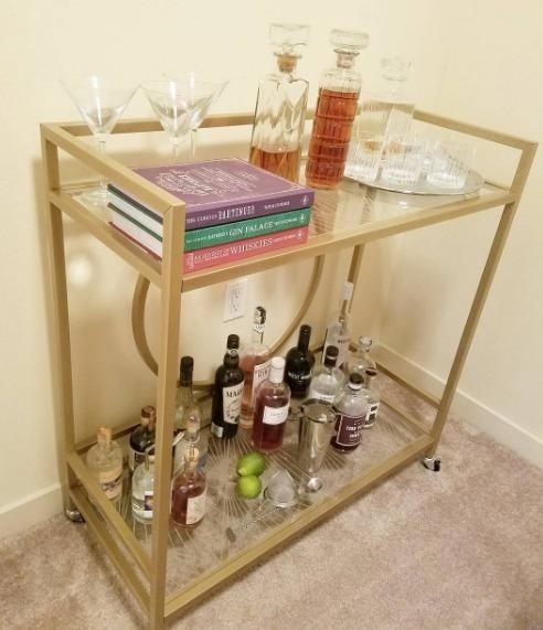 Reviewer pic of the bar cart with two glass-top shelves with a geometric pattern to them  and assorted alcohol bottles and glasses on each one. 